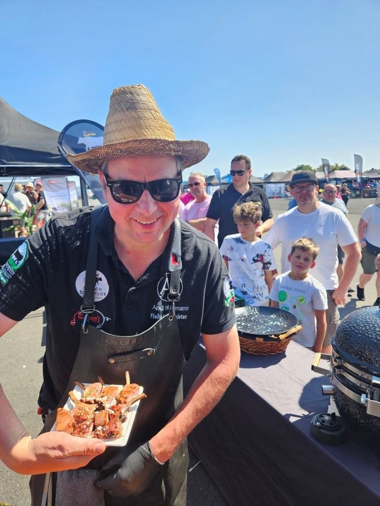 Eine lächelnde Person mit Strohhut und Sonnenbrille hält Essen auf einem Freiluftmarkt oder Festival.