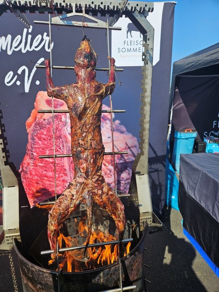 Ein ganzes, am Spieß über offenem Feuer gebratenes Tier an einem Marktstand im Freien, daneben werden Fleischstücke ausgestellt.