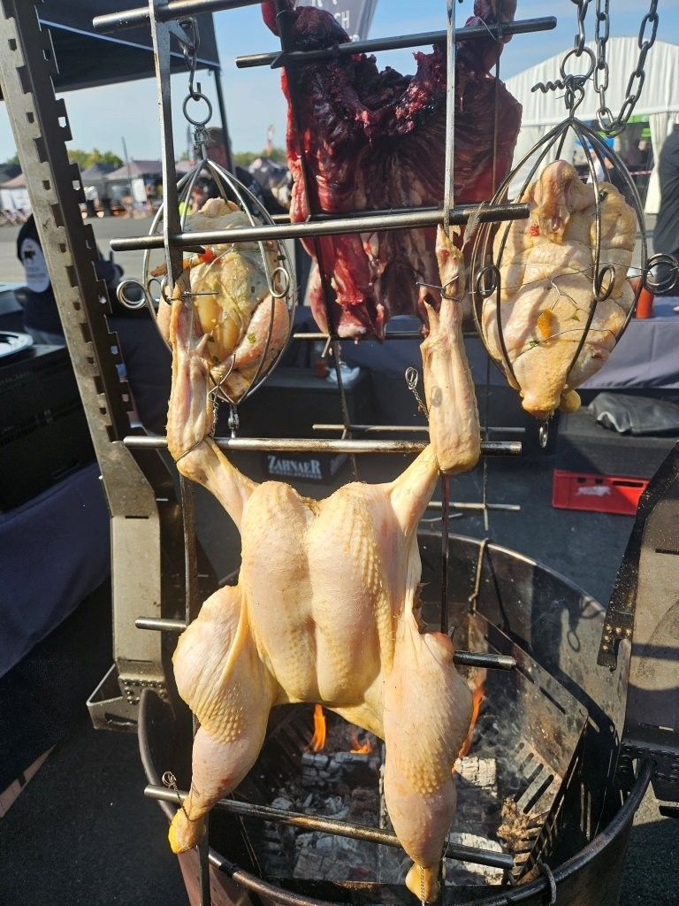 Drei Hühner braten im Freien auf einem Grill, im Hintergrund ist ein Fischerboot zu sehen.