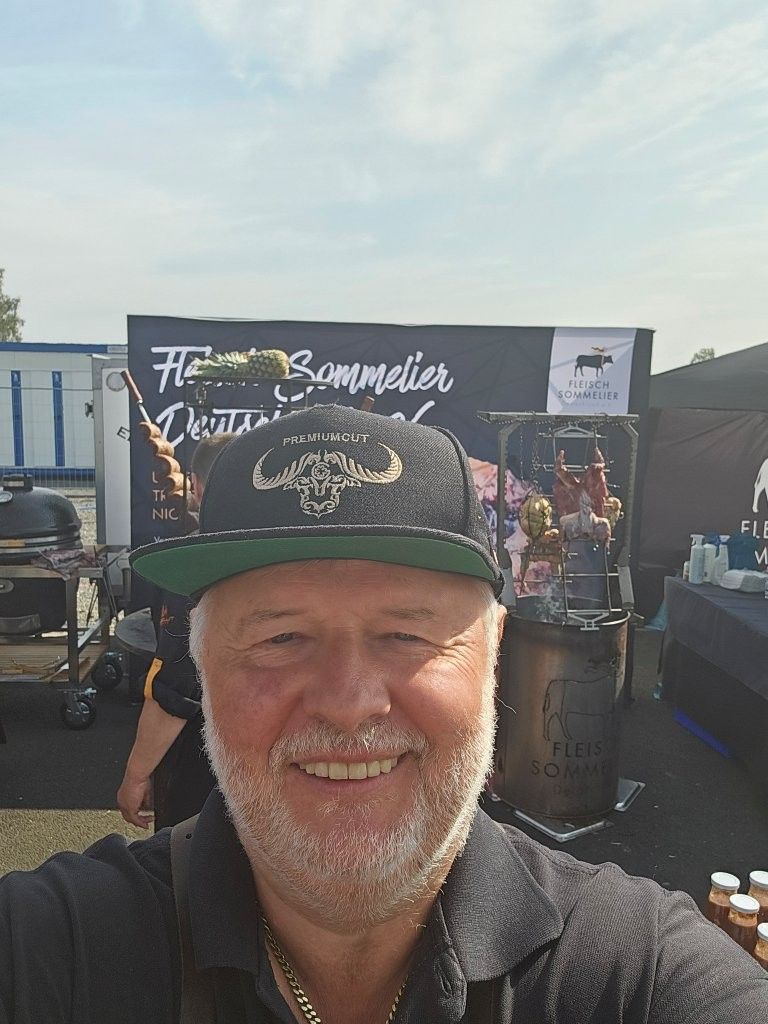 Ein lächelnder Mann mit schwarzer Kappe steht an einem Stand bei einer Open-Air-Veranstaltung; im Hintergrund ist ein Bandbanner zu sehen.