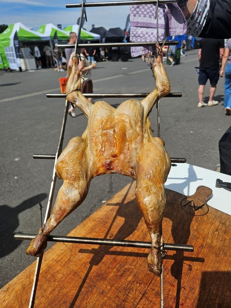 Ein ganzes Brathähnchen am Spieß auf einem Freiluftmarkt, im Hintergrund Zelte und Menschen.