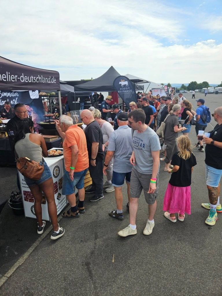 Menschenmenge steht an einem Essensstand im Freien auf einem Festivalgelände an, umgeben von Zelten und Besuchern in Freizeitkleidung.