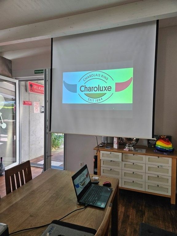 Konferenzraum mit einer Projektionsleinwand, auf der über einem Tisch mit einem Laptop das Cheluxe-Logo zu sehen ist.