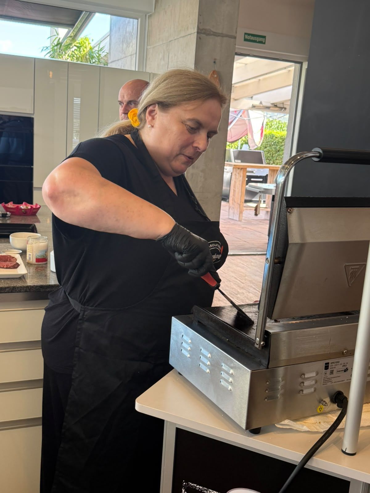 Eine Person grillt an einem Imbissstand im Freien und trägt dabei eine schwarze Schürze und Handschuhe.