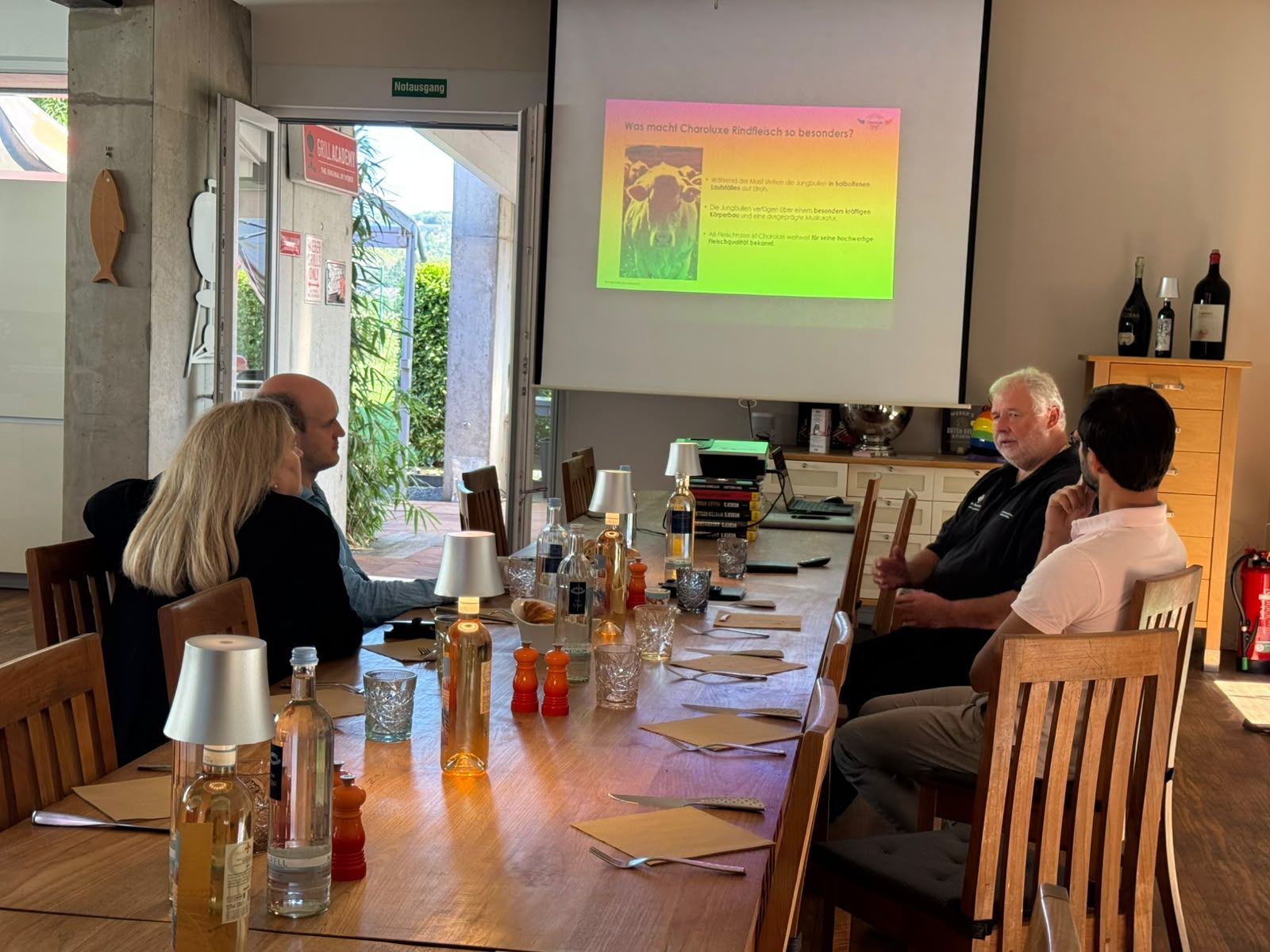 Menschen sitzen in einem Besprechungsraum an einem langen Tisch und sehen sich eine Präsentation auf einer Leinwand an.