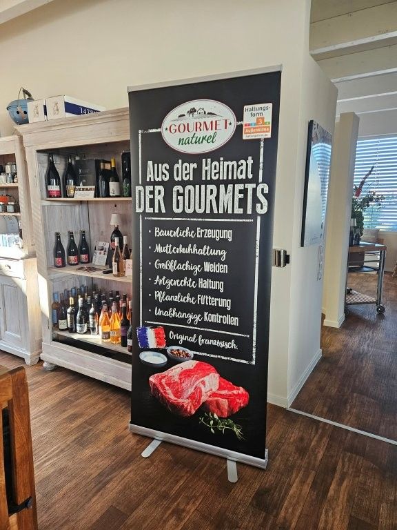 Schwarzes Restaurant-Schild mit der Aufschrift „Aus der Heimat der Gourmets“ neben einem gut gefüllten Regal in einem hellen Ladenlokal