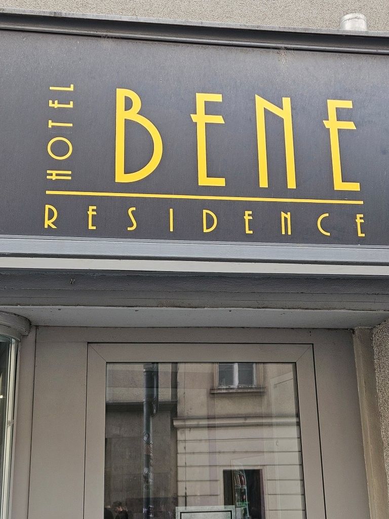 Schild mit der Aufschrift „BENE RESIDENCE“ in gelben Buchstaben über einem Gebäudeeingang.