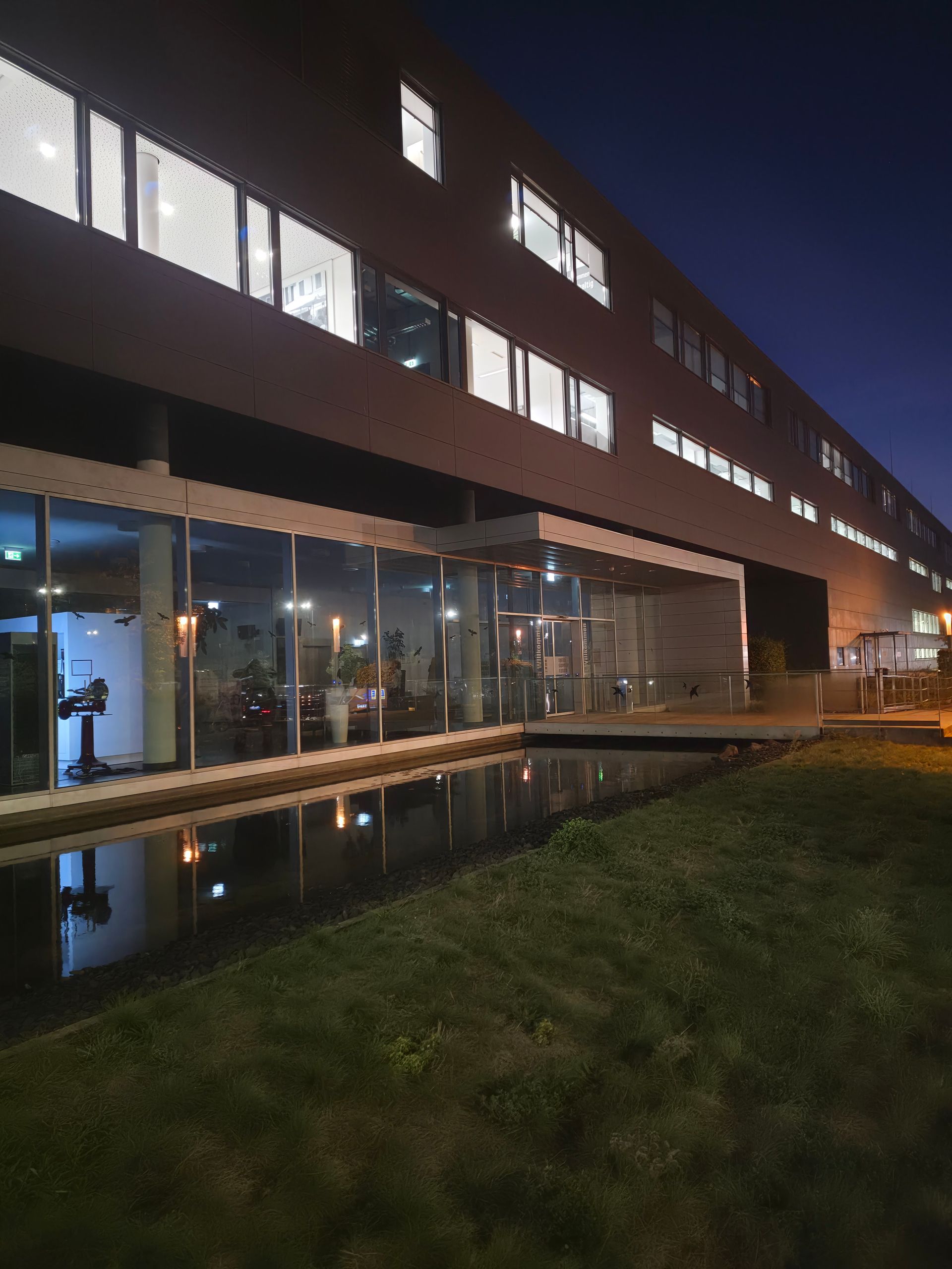 Modernes Bürogebäude bei Nacht mit beleuchteten Fenstern und einem gläsernen Eingang, der sich in einem Wasserspiel spiegelt
