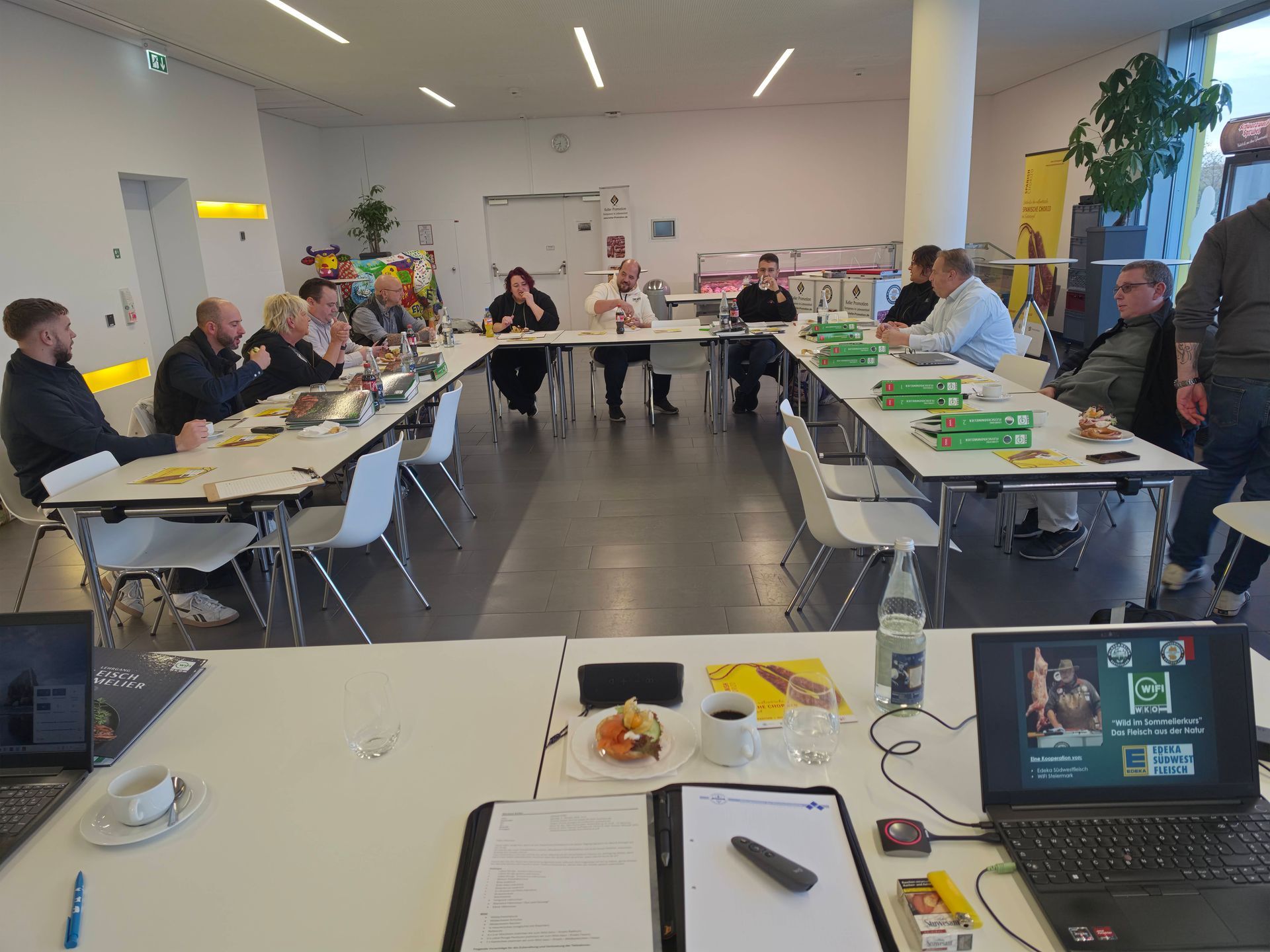 Menschen sitzen an U-förmigen Tischen in einem hellen Besprechungsraum, mit Laptops, Papieren und Snacks.