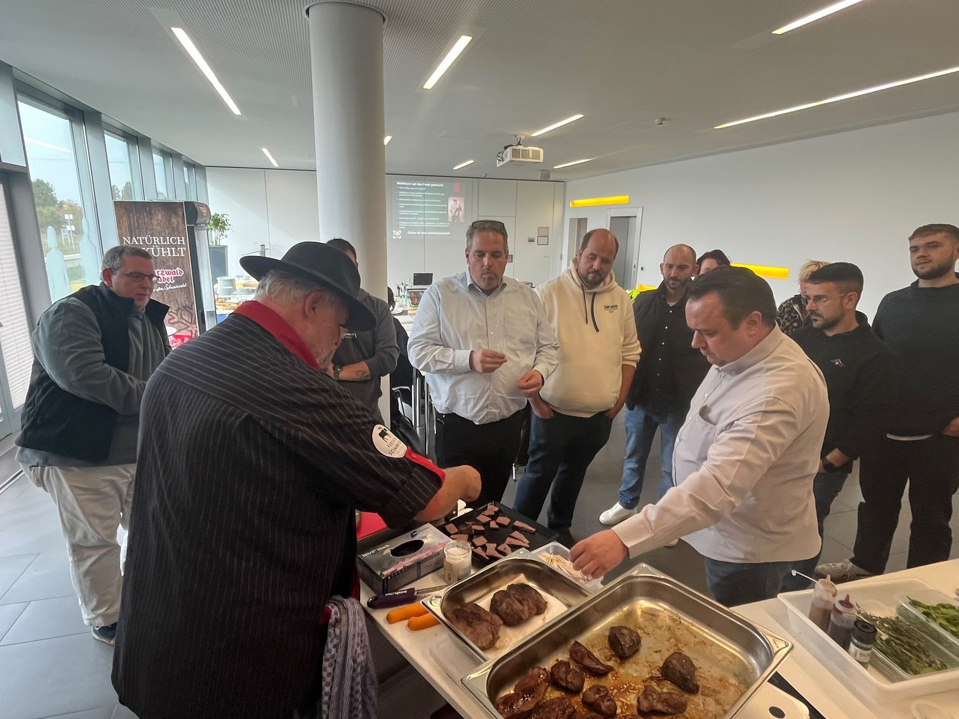 Menschen stehen an einem Buffet in einem hellen Veranstaltungsraum, nehmen sich Essen und unterhalten sich.