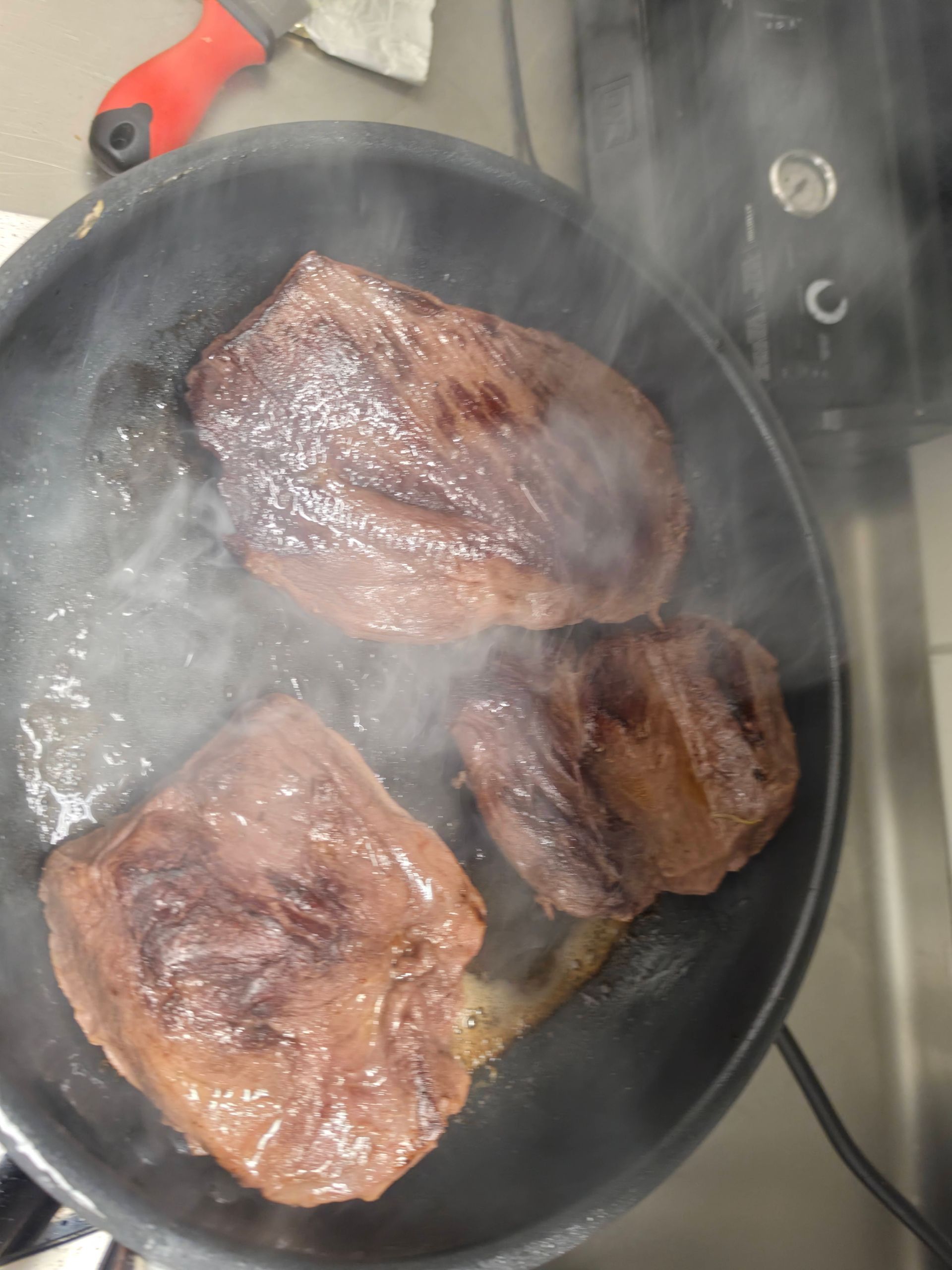 Drei Steaks braten in einer schwarzen Pfanne, während in einer Küche Dampf aufsteigt.