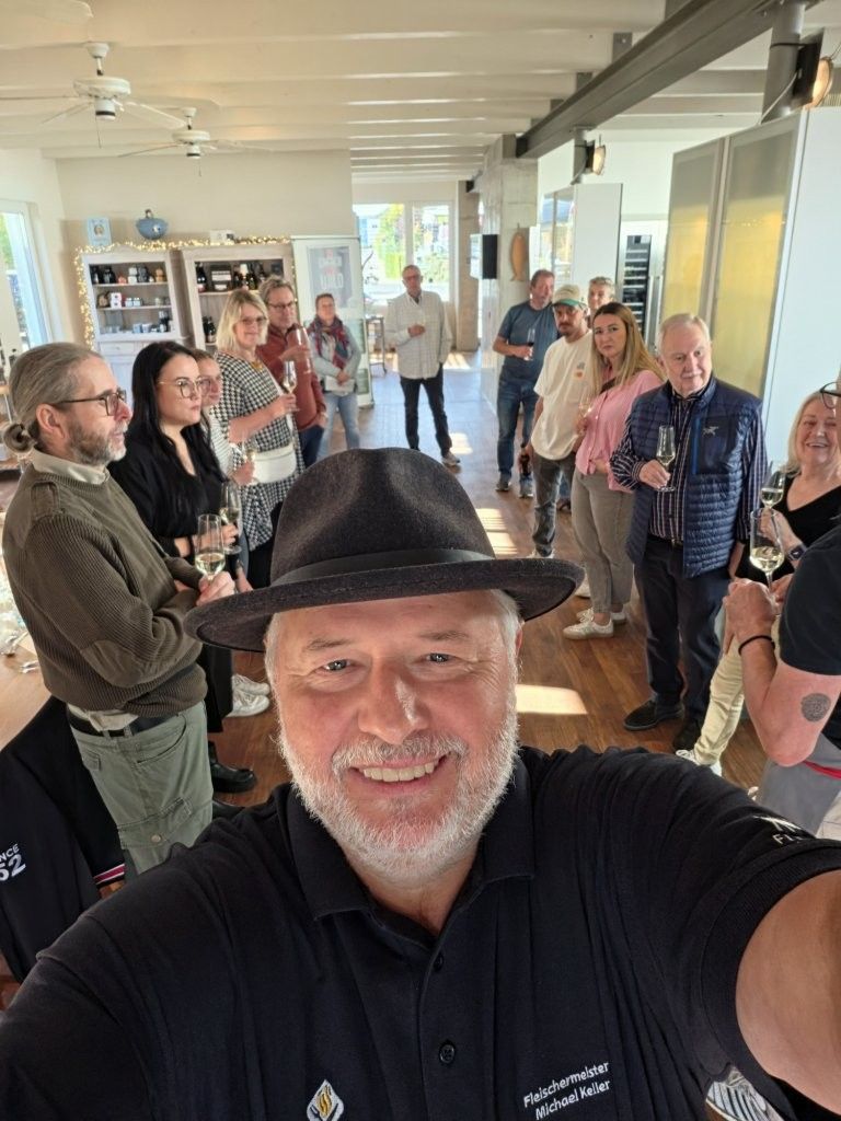 Eine Gruppe von Menschen steht in einem hellen Flur. Vorne macht ein lächelnder Mann mit schwarzem Hut ein Selfie.