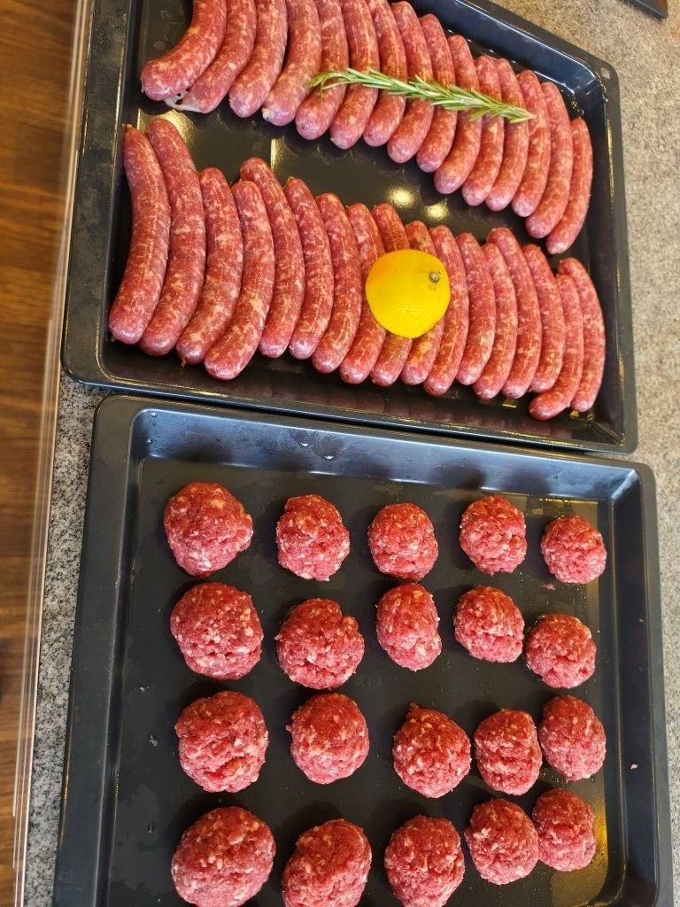 Rohe Würstchen und Burger-Patties auf schwarzen Tabletts angerichtet, garniert mit einer gelben Zitrone und Kräutern.