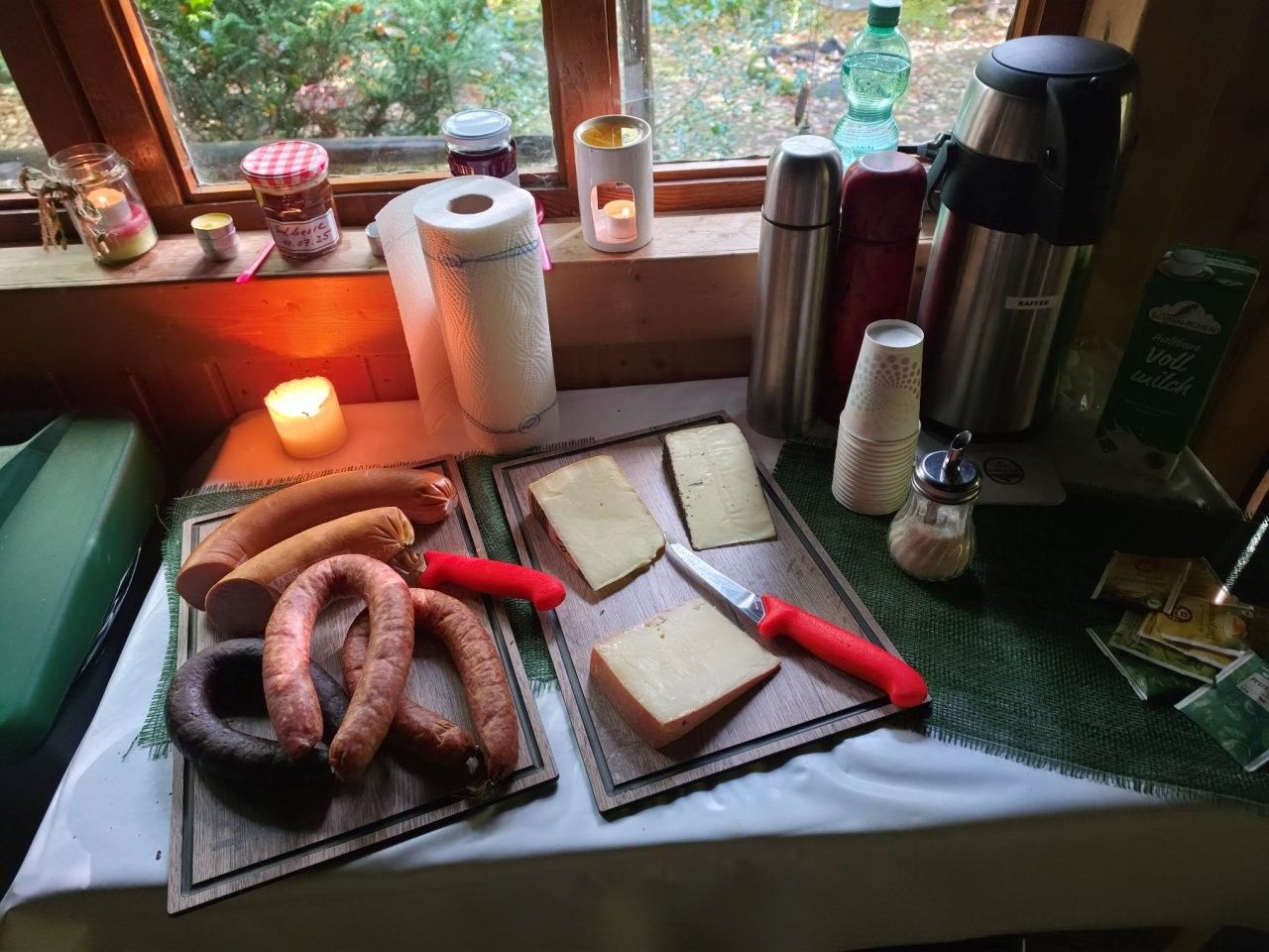Frühstücksaufstrich auf einer Fensterbank: Brotscheiben, Gebäck, ein Teelicht und Thermoskannen auf einer Tischdecke.
