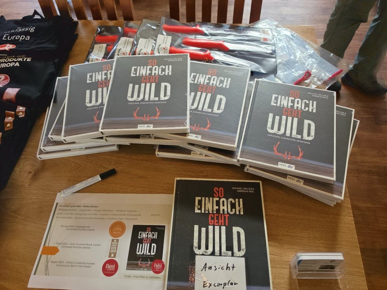 Ein Stapel „Einfach Wild“-Hefte und Broschüren liegt auf einem Tisch, mit schwarzem Einband und orangefarbenen Akzenten.