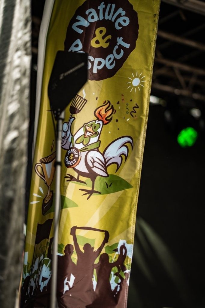 Gelbes Banner mit der Aufschrift „Natur & Respekt“ und einem Comic-Hahn sowie Silhouetten von Menschenmengen am unteren Rand.