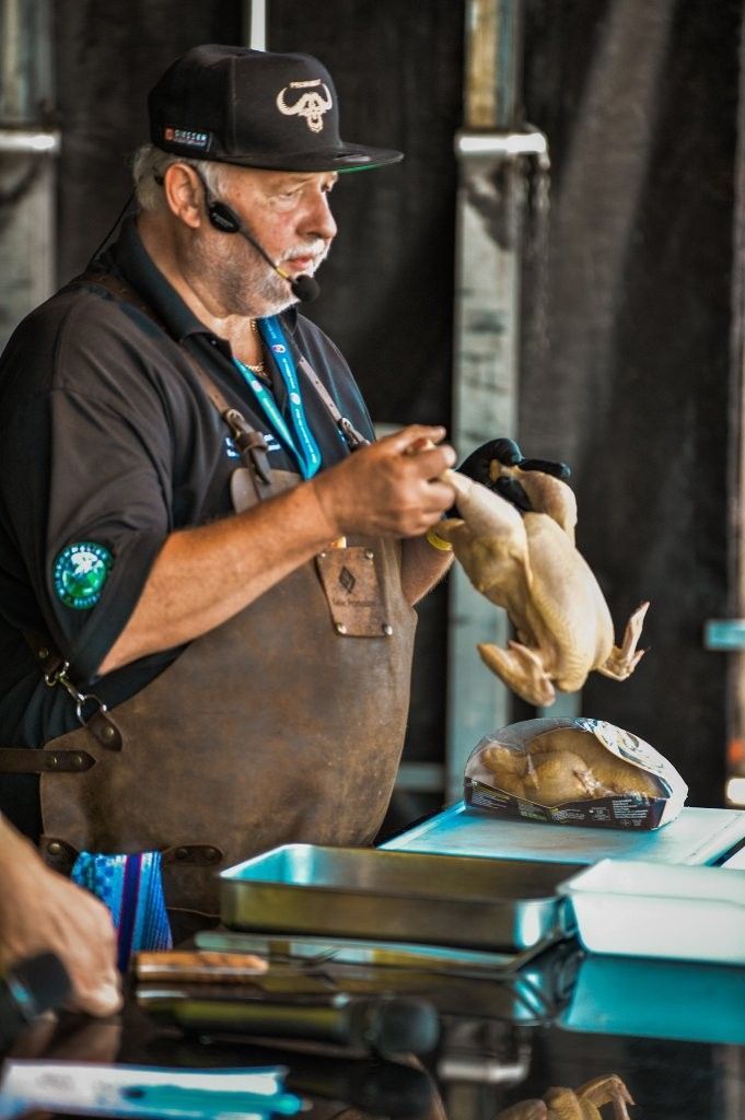 Ein Koch mit schwarzer Kochmütze und Schürze tranchiert Fleisch an einem Imbissstand und serviert ein Bratengericht über einer Theke.