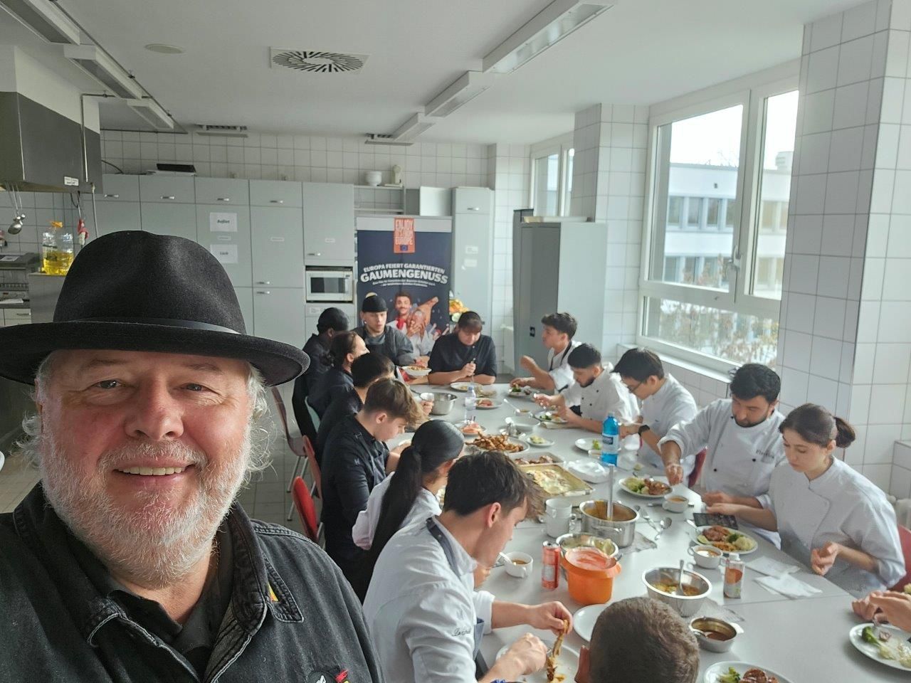 Ein Mann mit Hut macht ein Selfie mit einer Gruppe von Menschen, die an Tischen sitzen und Fleisch verarbeiten