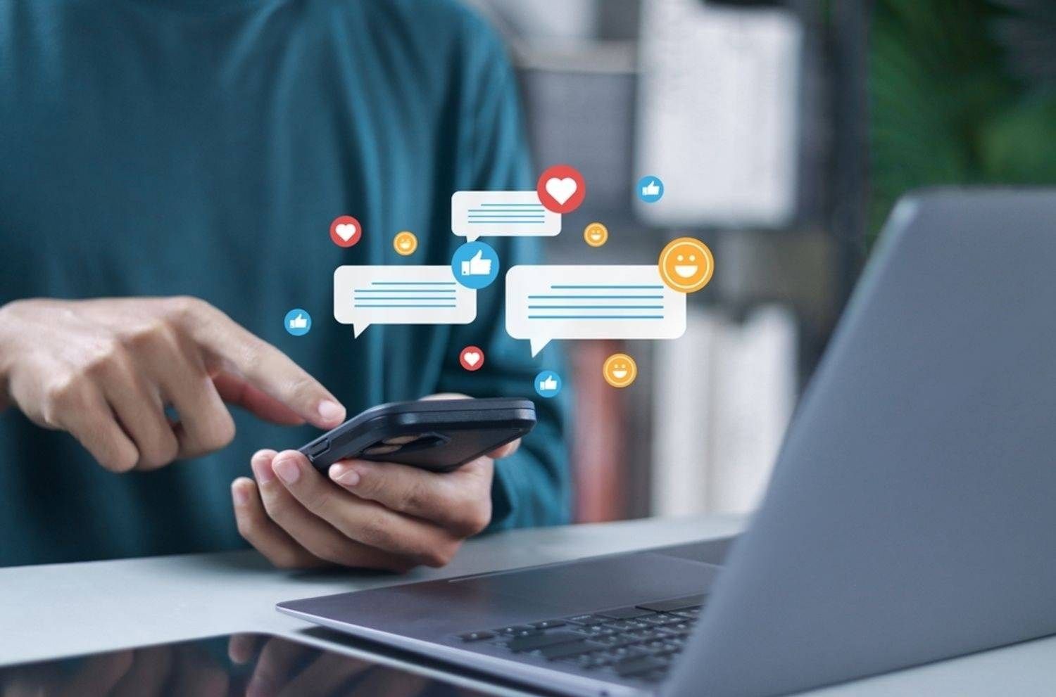 Person, die ein Smartphone mit schwebenden Social-Media-Symbolen in der Nähe eines Laptops benutzt.