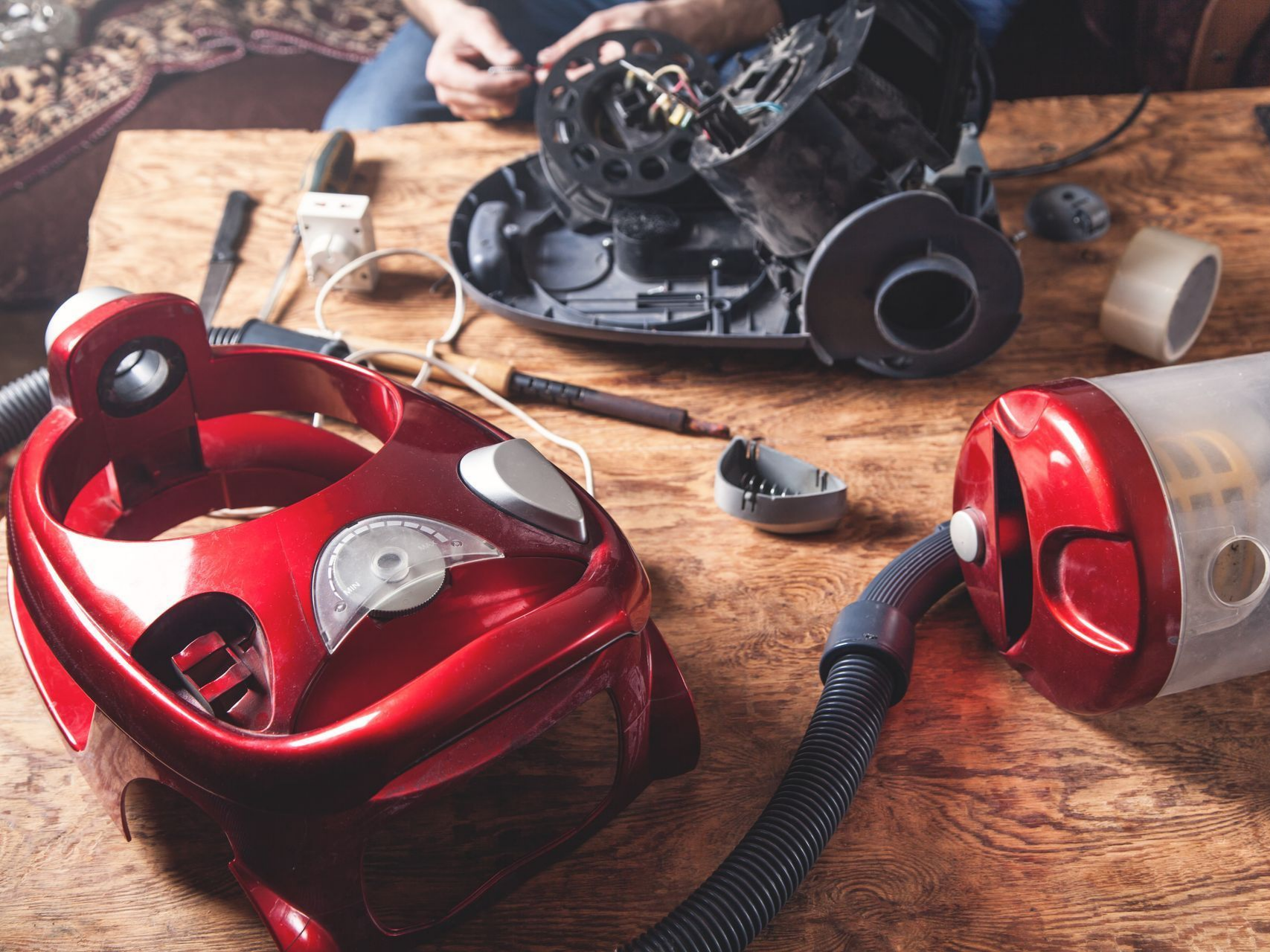 Aspirateur démonté sur une table en bois, avec ses pièces et outils. Une personne est visible en arrière-plan.