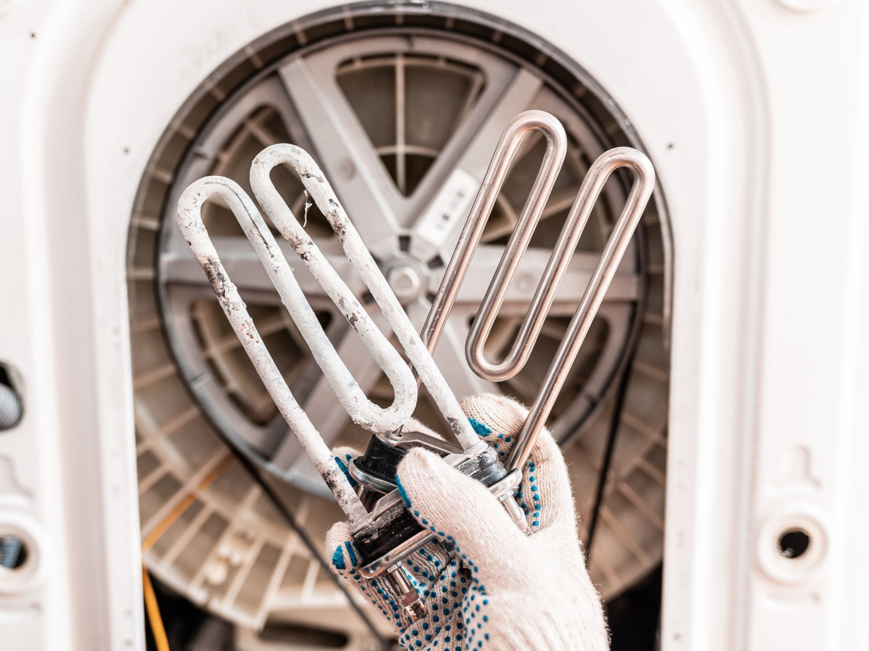 Une main tenant un élément chauffant corrodé provenant d'une machine à laver, avec les mécanismes internes de la machine en arrière-plan.