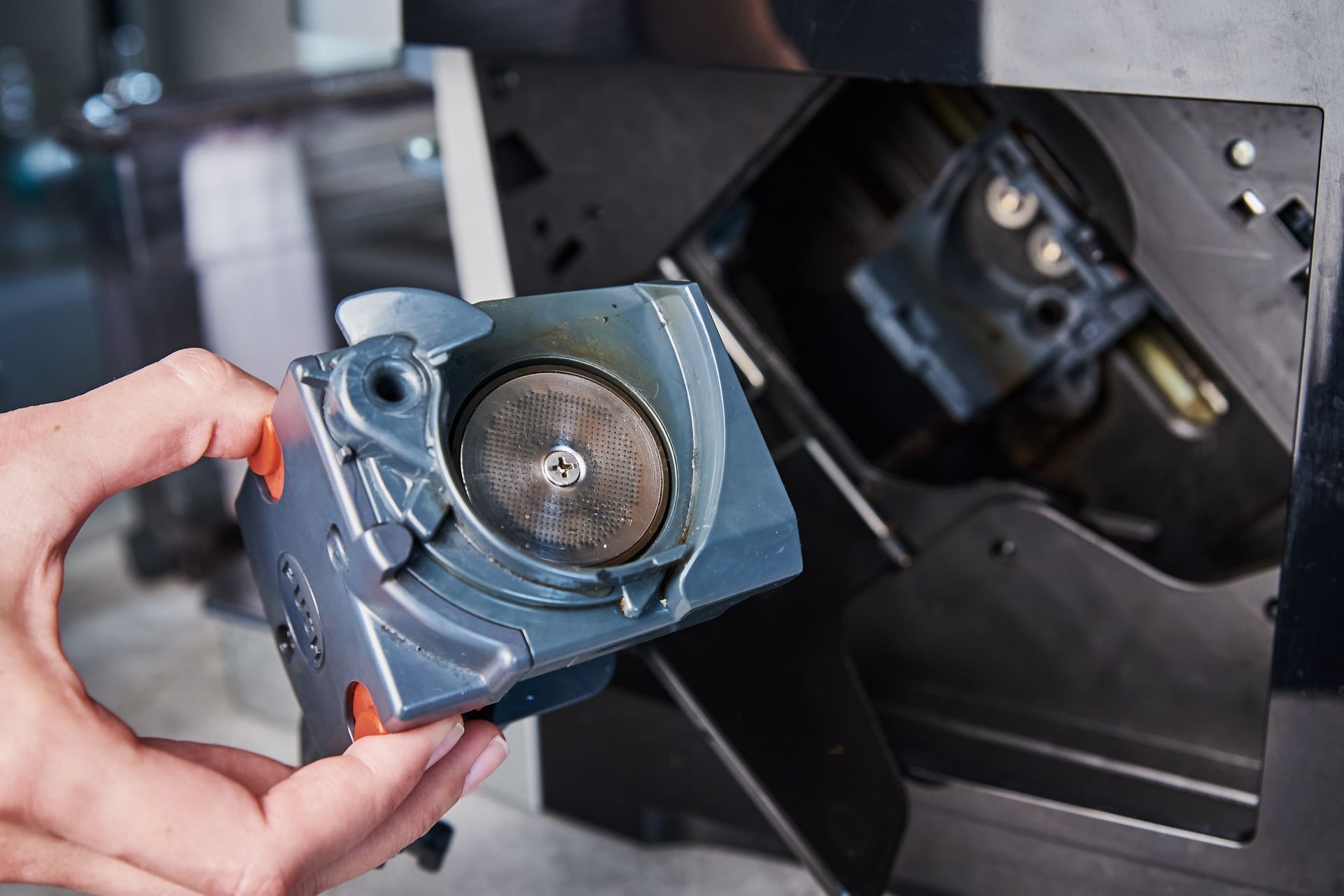 Une main tenant un composant gris d'une machine à café, révélant l'intérieur ; un compartiment noir de la machine est visible.