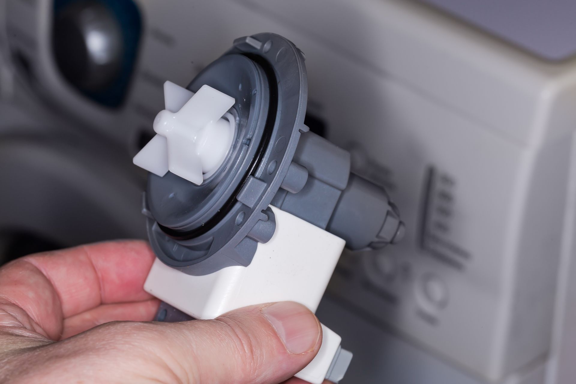 Main tenant une pompe de vidange de machine à laver, turbine en plastique blanc, boîtier gris, machine à laver en arrière-plan.
