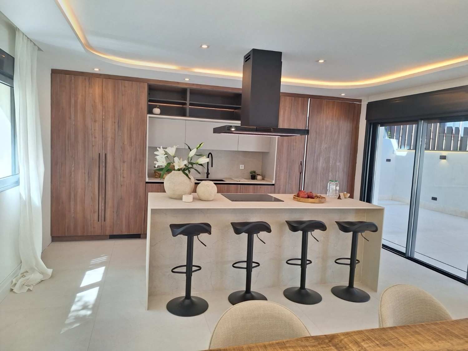 Cocina moderna con isla, taburetes negros, armarios de madera y puertas correderas de cristal que dan a un patio.