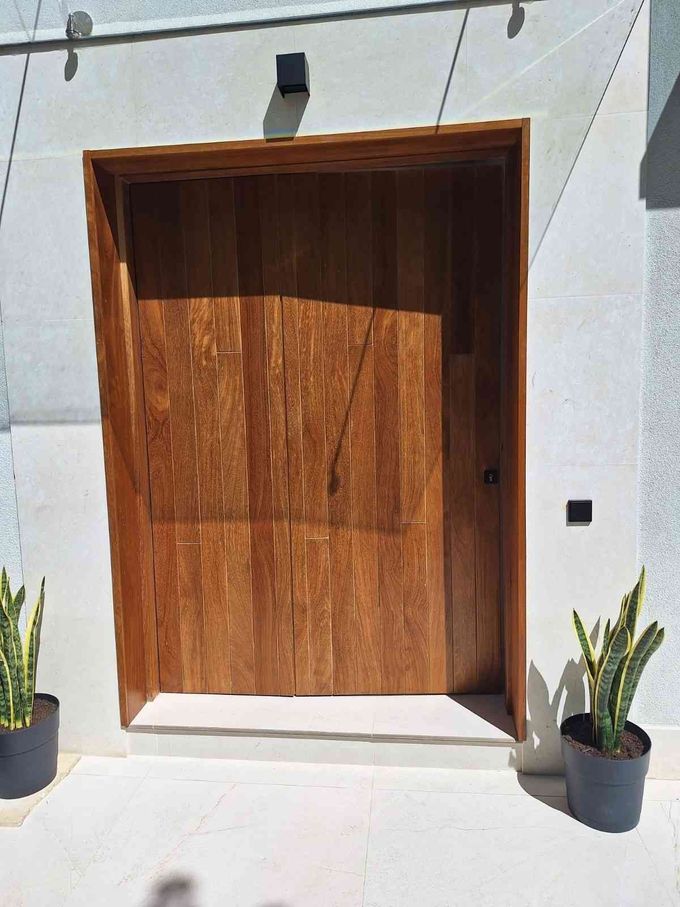 Puerta de entrada moderna de madera entre dos macetas en una pared exterior blanca.
