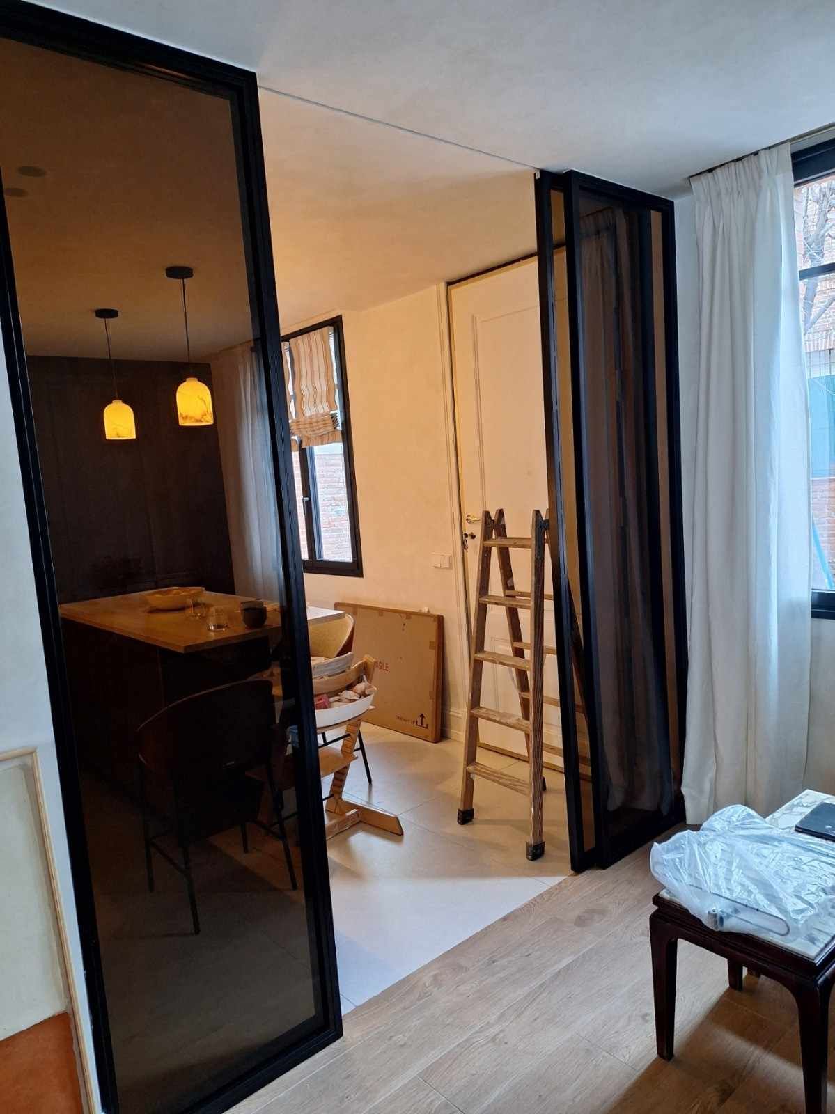Interior de un apartamento moderno con tabiques de cristal, una escalera y un comedor junto a una ventana luminosa.