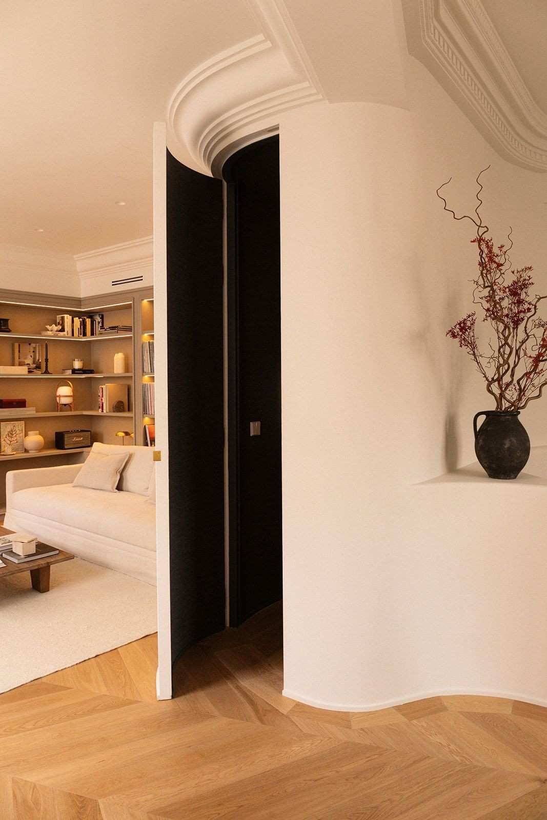 Elegante habitación con puerta curva blanca, suelo de madera, sofá beige y jarrón con ramas secas en una estantería.