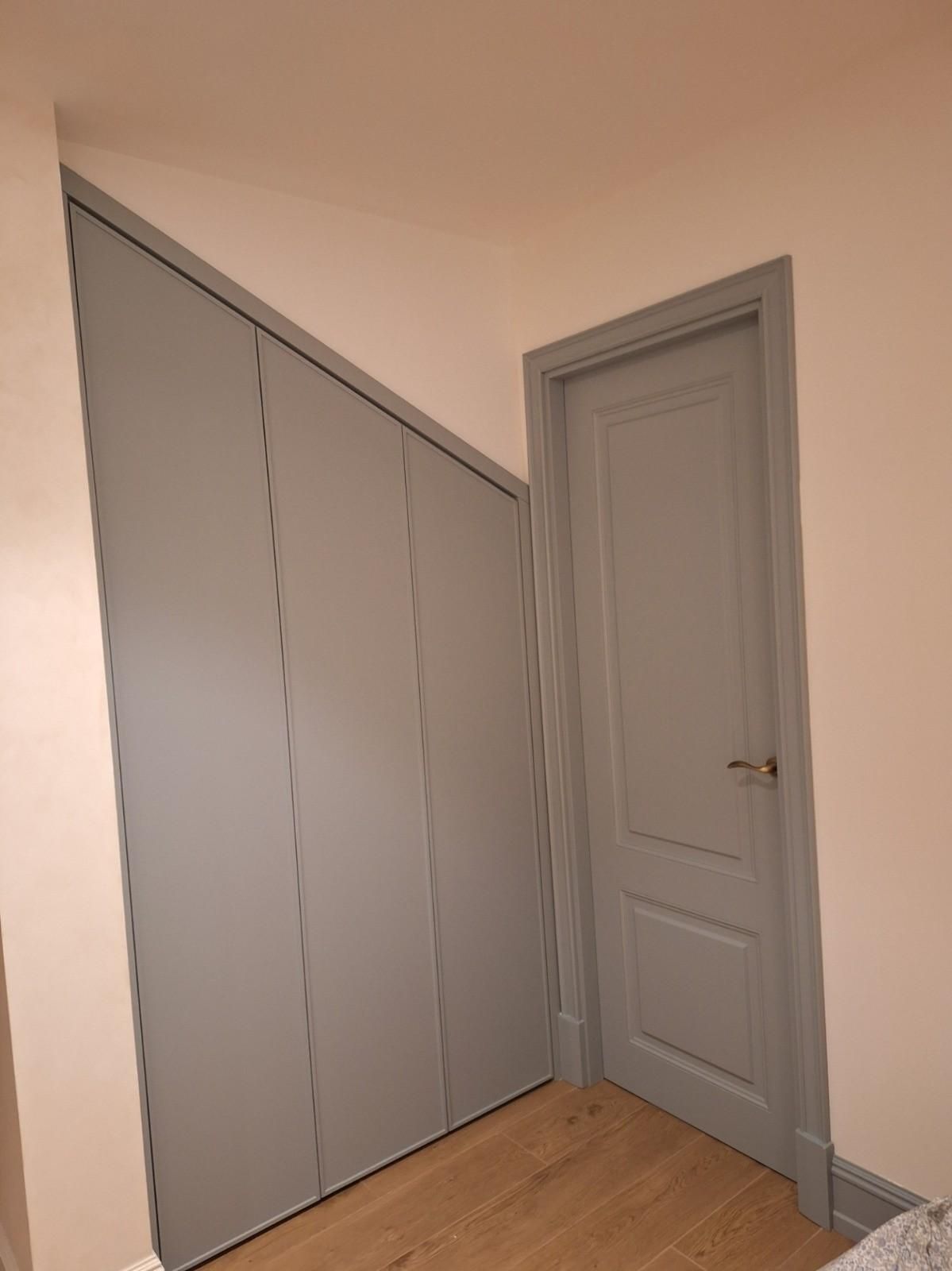 Armario empotrado gris bajo un techo inclinado, junto a una puerta con paneles grises, en una habitación de color beige.