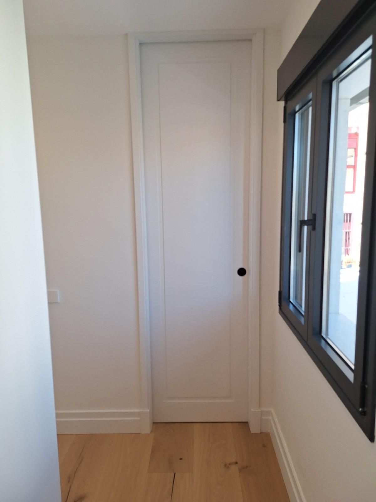 Pasillo estrecho de color blanco con una puerta cerrada, suelo de madera clara y una ventana a la derecha.