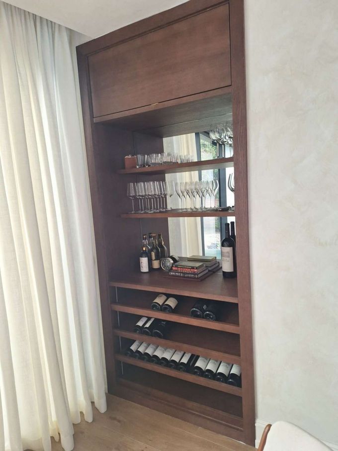 Estantería de madera empotrada con libros, botellas y ropa doblada junto a cortinas blancas.