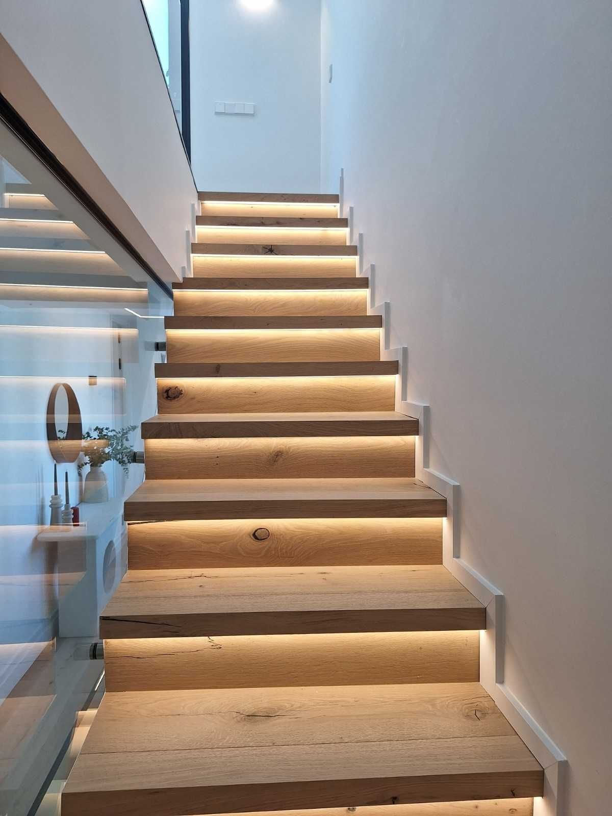 Escalera interior estrecha con paredes blancas, escalones de madera y una barandilla de cristal a la izquierda.