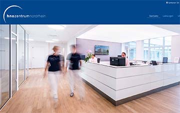 Empfangsbereich einer modernen medizinischen Klinik; verschwommene Figuren, strahlend weißer Schreibtisch, blaue Akzente.