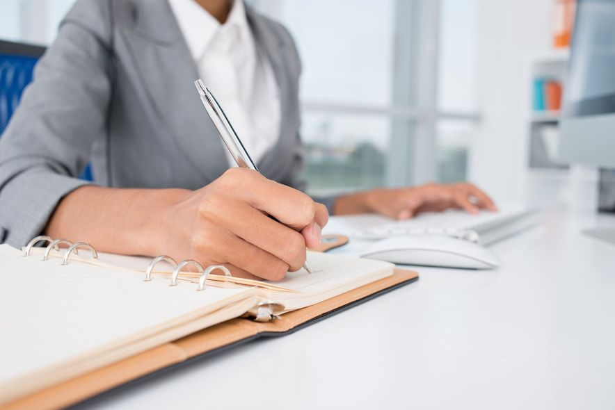 Image portrait d'une femme qui écrit sur un agenda papier devant un clavier d'ordinateur