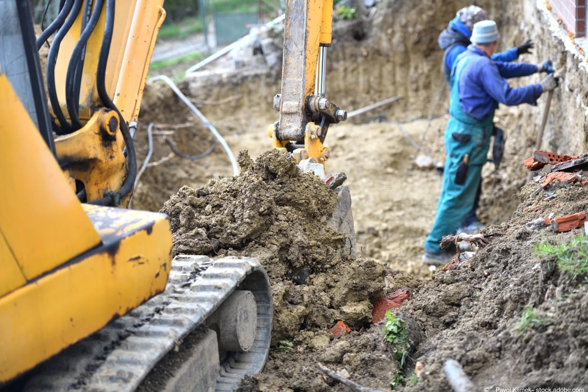 Ein kleiner Bagger schaufelt, zwei Arbeiter stehen ein paar Meter weiter und schaufeln ebenfalls