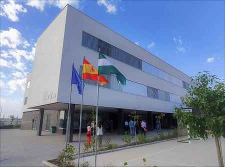 Edificio de oficinas blanco moderno con banderas españolas y de la UE, gente en la entrada, bajo un cielo azul.