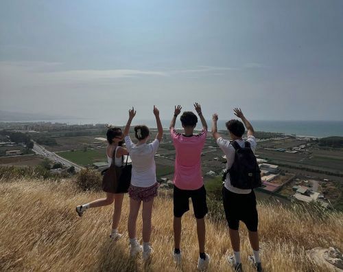 Cuatro personas están de pie en una colina, con los brazos levantados hacia el cielo con vistas al océano y al paisaje.