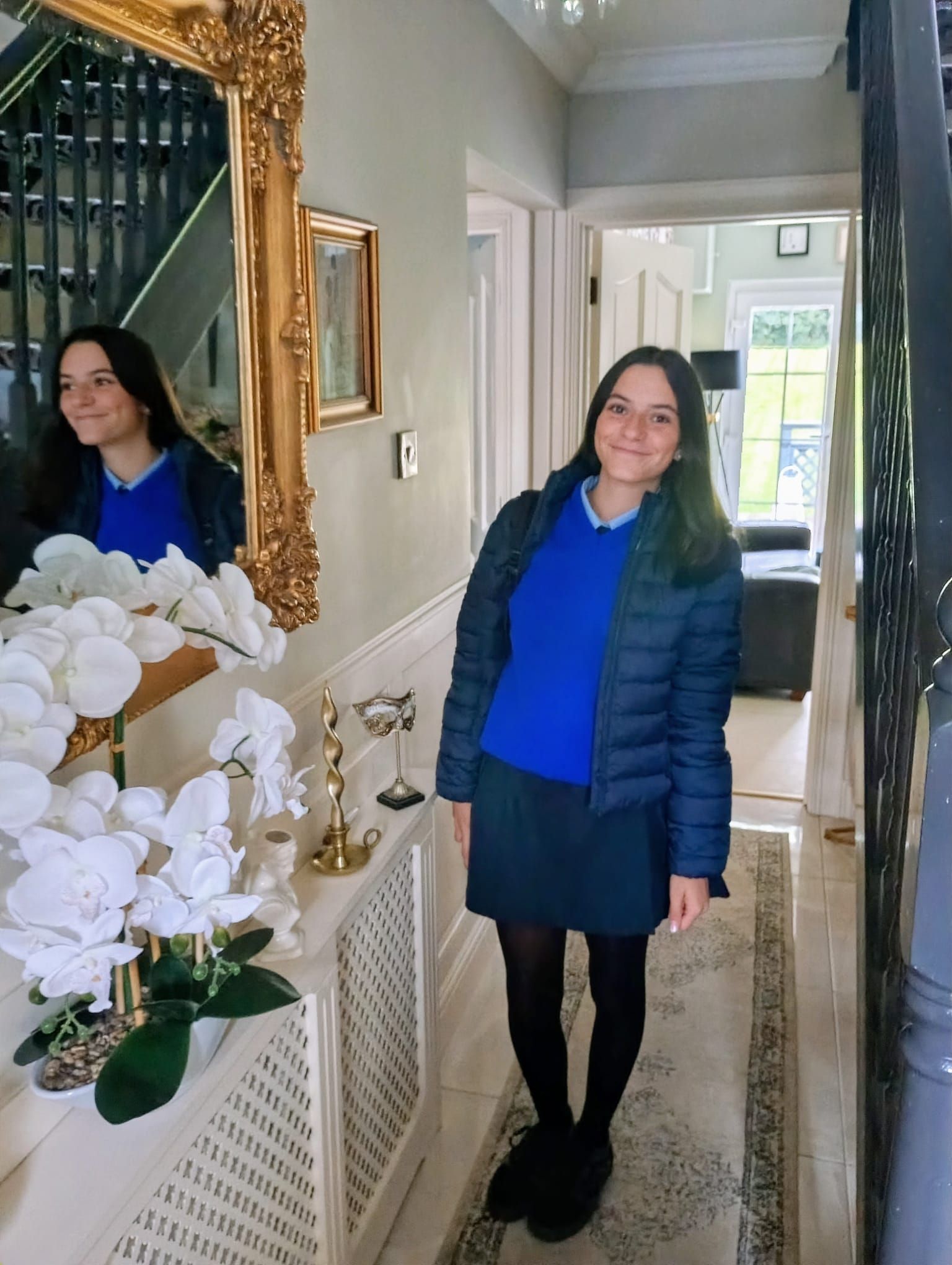 Mujer con suéter azul de pie en un pasillo luminoso junto a una escalera y un espejo.