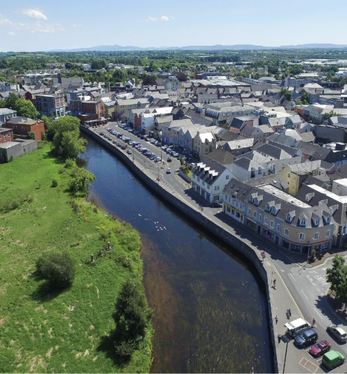 Vista aérea de la ciudad junto a un río, campos verdes y edificios, día soleado.