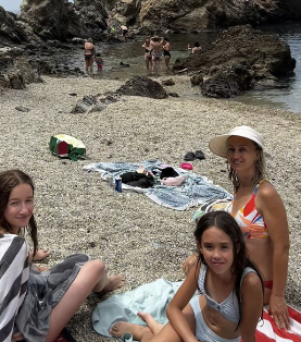 Familia relajándose en una playa de guijarros; mujer con sombrero y bikini; niñas y otras personas en el agua.
