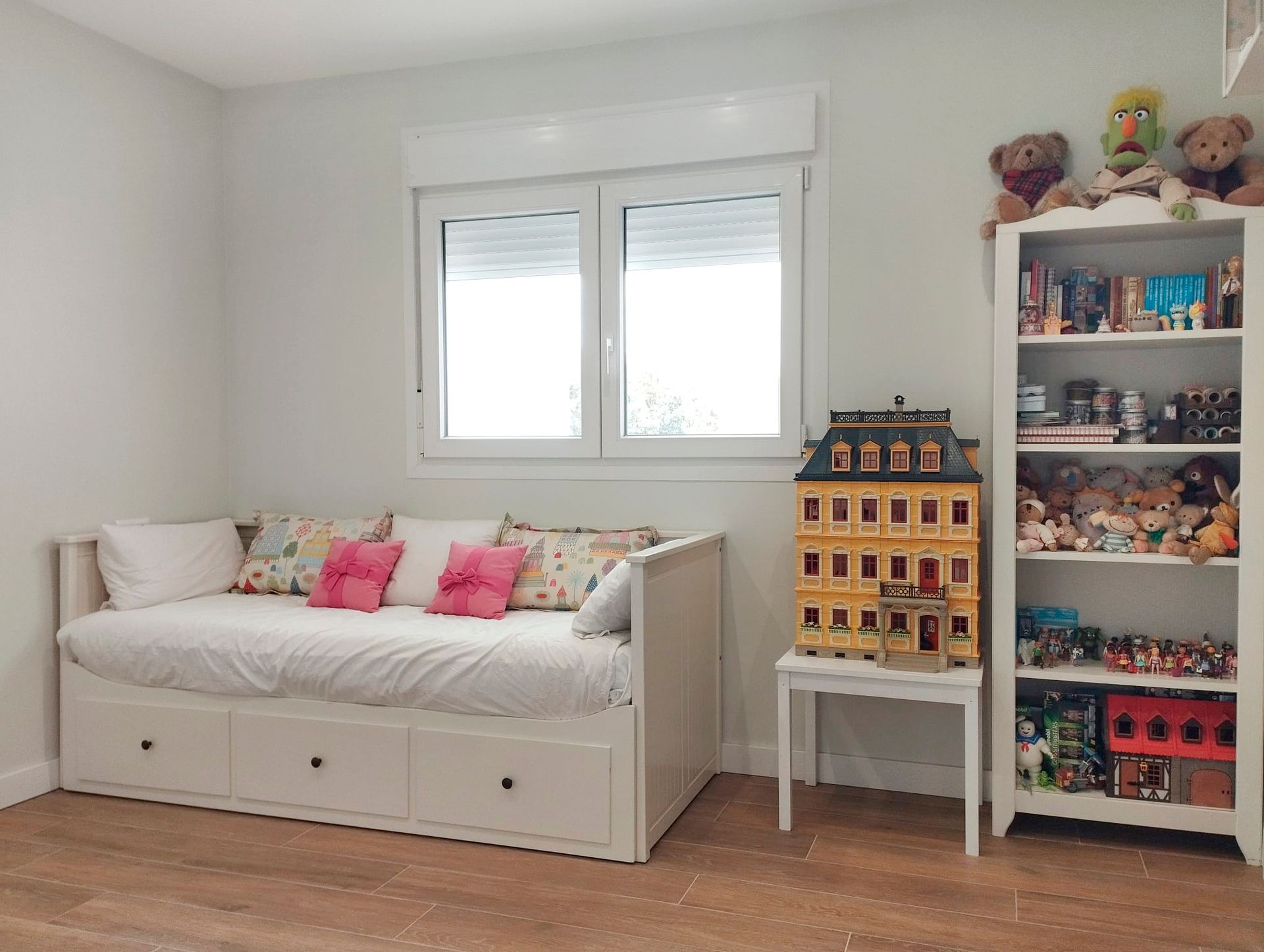 Un dormitorio infantil con sofá-cama, ventana y estantería llena de juguetes.
