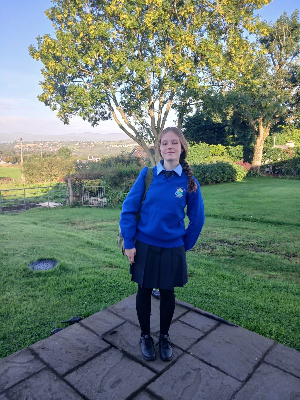Estudiante con uniforme escolar azul de pie en un patio pavimentado en un jardín soleado.