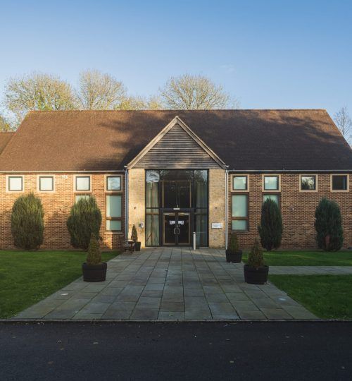 Edificio de ladrillo marrón con entrada de vidrio, camino de piedra y arbustos podados en el frente.