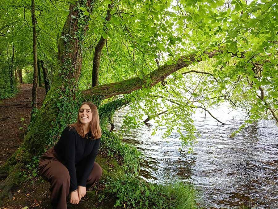 Mujer sentada junto a un árbol inclinado al lado de un río, sonriendo en una zona boscosa verde.