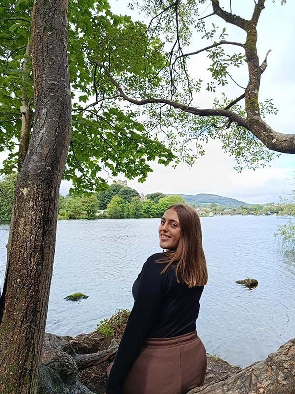 Mujer de pie junto a un lago bajo los árboles, sonriendo a la cámara con el agua y las colinas de fondo.