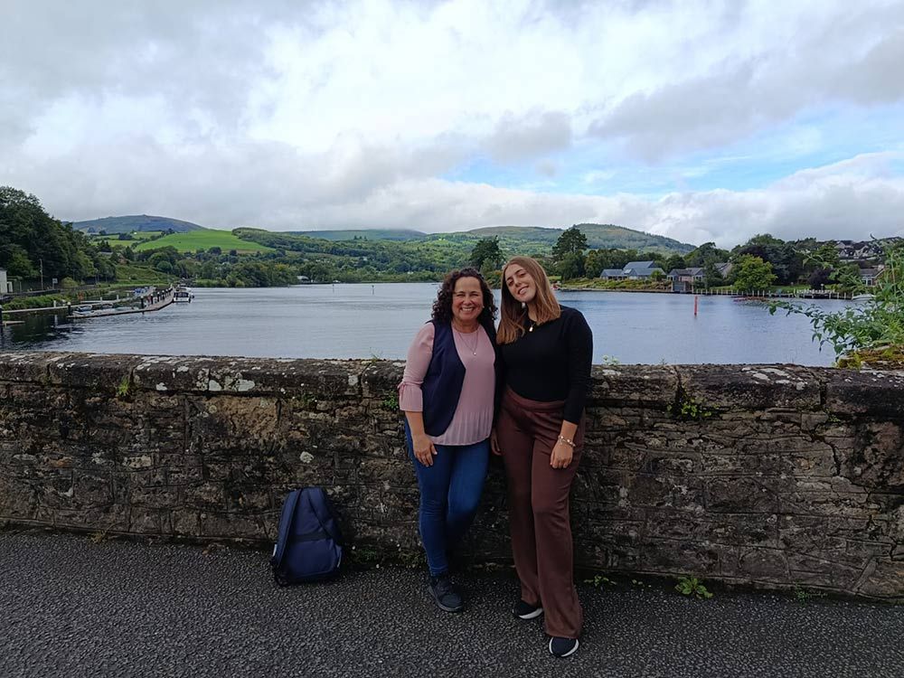 Dos personas posando junto a un muro de piedra con vistas a un lago y verdes colinas bajo un cielo nublado.