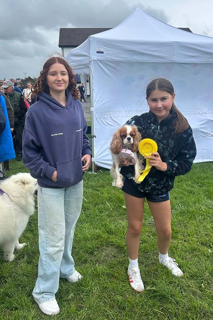 Dos niñas con perros en brazos al aire libre, cerca de una tienda de campaña blanca; uno de ellos llevaba una cinta amarilla y una flor.